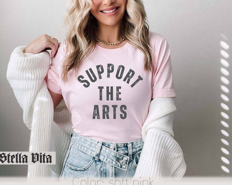 May include: A pink t-shirt with the text "Support the Arts" in gray lettering. The shirt is being worn by a person with long blonde hair.