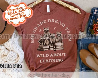 Puede incluir: Una camiseta de color arcilla con un gráfico de mapaches sentados en una pila de libros. El texto "3rd Grade Dream Team" está en un círculo alrededor de los mapaches. El texto "Wild About Learning" está debajo de los mapaches.