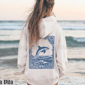 May include: A white hoodie with a blue and white graphic of an orca jumping out of the water surrounded by flowers. The graphic is on the back of the hoodie.