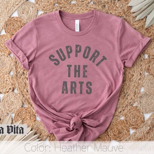 May include: A heather mauve t-shirt with the text "Support the Arts" in a black, distressed font.