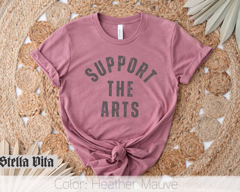 May include: A heather mauve t-shirt with the text "Support the Arts" in a black, distressed font.