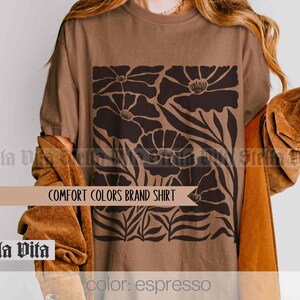 Puede incluir: Una camiseta marr&oacute;n con un dise&ntilde;o floral en negro. El dise&ntilde;o presenta flores estilizadas con p&eacute;talos grandes y hojas. El texto "COMFORT COLORS BRAND SHIRT" est&aacute; impreso en la camiseta. El color de la camiseta se describe como "espresso".