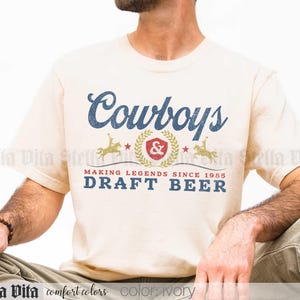 May include: Ivory-colored t-shirt with the word "Cowboys" in blue script. Below, a graphic with a cowboy on a horse, a red emblem, and the text "MAKING LEGENDS SINCE 1985 DRAFT BEER" in blue. The shirt is a comfort colors brand.