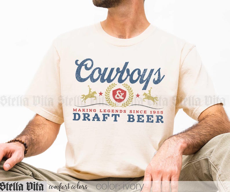 May include: Ivory-colored t-shirt with the word "Cowboys" in blue script. Below, a graphic with a cowboy on a horse, a red emblem, and the text "MAKING LEGENDS SINCE 1985 DRAFT BEER" in blue. The shirt is a comfort colors brand.