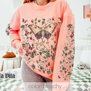May include: Peach colored sweatshirt with a large butterfly and floral design. The butterfly is a light brown color with black and white markings. The flowers are pink and white.