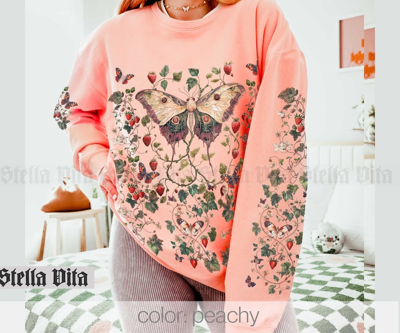 May include: Peach colored sweatshirt with a large butterfly and floral design. The butterfly is a light brown color with black and white markings. The flowers are pink and white.