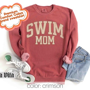 Puede incluir: Sudadera de color rojo carmes&iacute; con el texto "SWIM MOM" en una fuente de estilo vintage. La sudadera est&aacute; hecha de una tela suave y c&oacute;moda y tiene un corte holgado.