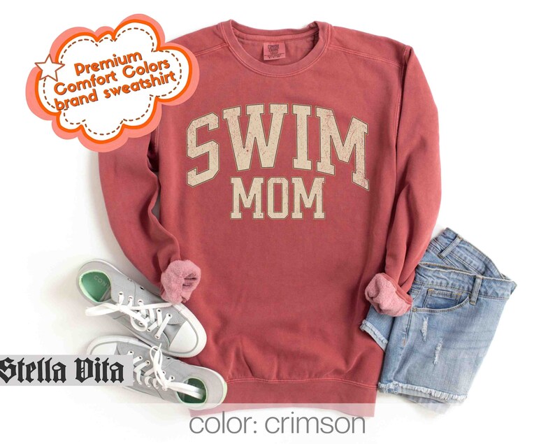 Puede incluir: Sudadera de color rojo carmes&iacute; con el texto "SWIM MOM" en una fuente de estilo vintage. La sudadera est&aacute; hecha de una tela suave y c&oacute;moda y tiene un corte holgado.