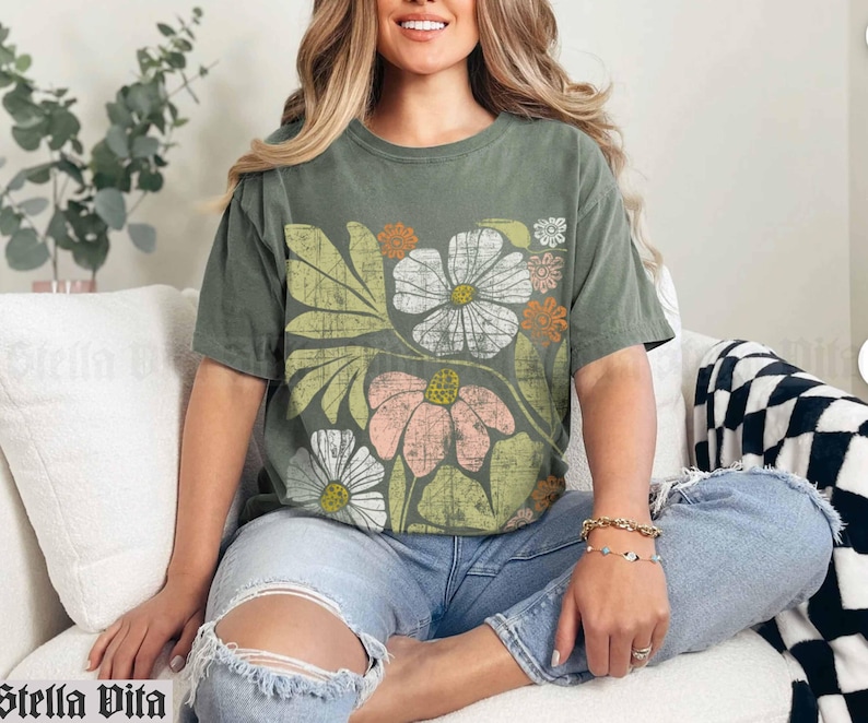 May include: A green t-shirt with a floral print featuring pink, white, and yellow flowers. The shirt has a relaxed fit and a crew neck.