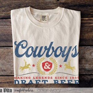 May include: Ivory-colored t-shirt with the word "Cowboys" in blue script. Below, a design includes a shield, stars, and the text "MAKING LEGENDS SINCE 1985 DRAFT BEER." Two figures on horseback flank the shield. The shirt is made by Comfort Colors.