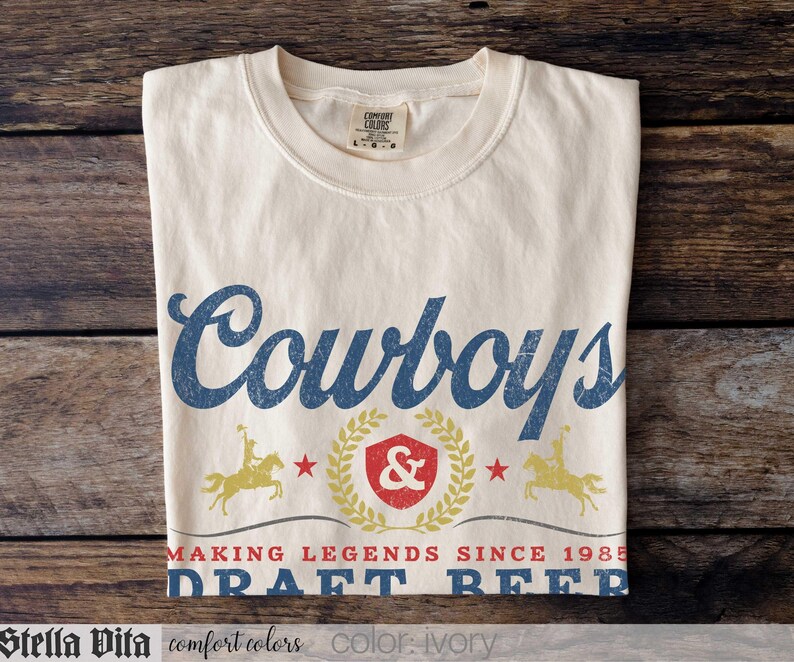 May include: Ivory-colored t-shirt with the word "Cowboys" in blue script. Below, a design includes a shield, stars, and the text "MAKING LEGENDS SINCE 1985 DRAFT BEER." Two figures on horseback flank the shield. The shirt is made by Comfort Colors.
