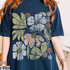 May include: A navy blue t-shirt with a floral print featuring large, stylized flowers in shades of pink, white, and brown. The flowers are surrounded by green leaves and have a vintage, retro aesthetic.