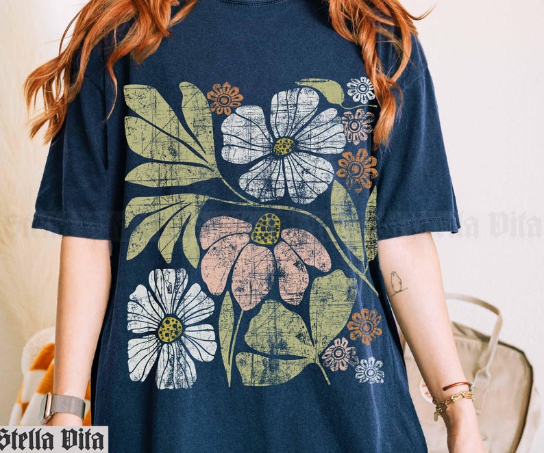 May include: A navy blue t-shirt with a floral print featuring large, stylized flowers in shades of pink, white, and brown. The flowers are surrounded by green leaves and have a vintage, retro aesthetic.
