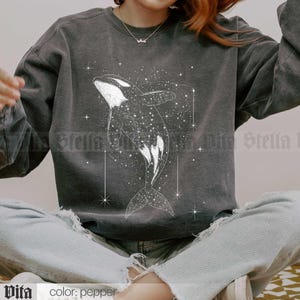 May include: A gray sweatshirt with a white graphic of an orca whale swimming through a field of stars. The text "Stella Vita" is printed on the bottom left corner of the sweatshirt.