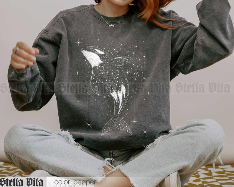 May include: A gray sweatshirt with a white graphic of an orca whale swimming through a field of stars. The text "Stella Vita" is printed on the bottom left corner of the sweatshirt.