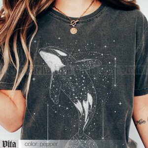 May include: A dark gray t-shirt with a white graphic of an orca whale swimming through a field of stars. The text "Stella Vita" is printed on the bottom left corner of the shirt. The text "color: pepper" is printed on the bottom right corner of the shirt.
