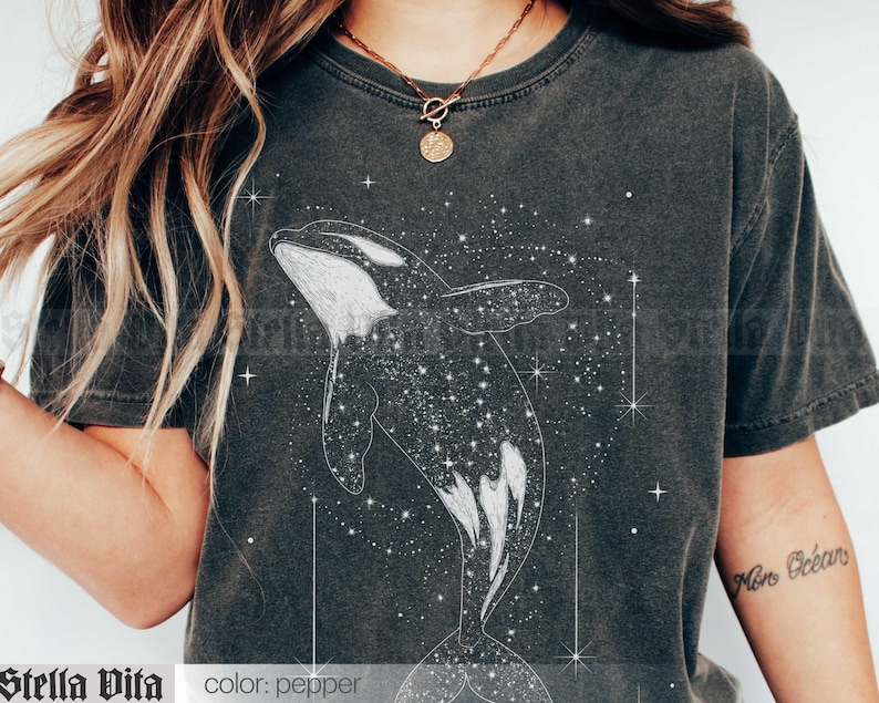 May include: A dark gray t-shirt with a white graphic of an orca whale swimming through a field of stars. The text "Stella Vita" is printed on the bottom left corner of the shirt. The text "color: pepper" is printed on the bottom right corner of the shirt.
