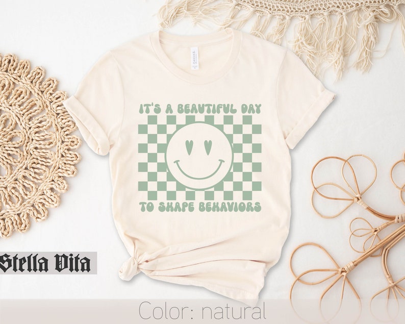 May include: A natural colored t-shirt with a green checkered pattern and a green smiley face with heart eyes. The text "It's a beautiful day to shape behaviors" is printed on the shirt.