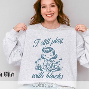 K&ouml;nnte beinhalten: Ein grauer Pullover mit dem Text "I still play with blocks" und einer Cartoon-Illustration eines M&auml;dchens, das mit Bl&ouml;cken spielt. Der Text "color: ash" befindet sich am unteren Rand des Pullovers.