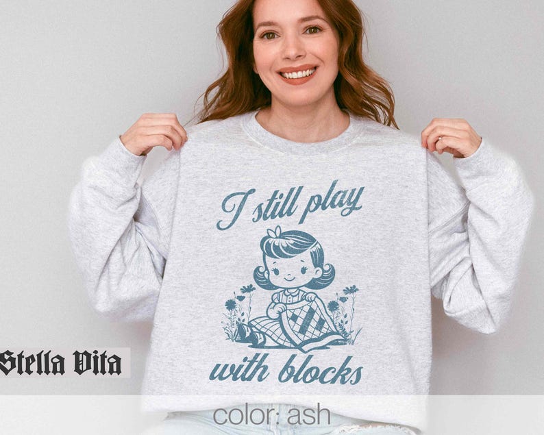 K&ouml;nnte beinhalten: Ein grauer Pullover mit dem Text "I still play with blocks" und einer Cartoon-Illustration eines M&auml;dchens, das mit Bl&ouml;cken spielt. Der Text "color: ash" befindet sich am unteren Rand des Pullovers.