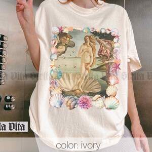 Puede incluir: Camiseta de color marfil con un estampado gr&aacute;fico de una pintura de estilo renacentista de una mujer de pie sobre una concha, rodeada de un borde floral de conchas. El texto "color: ivory" est&aacute; impreso en la parte inferior de la camiseta.