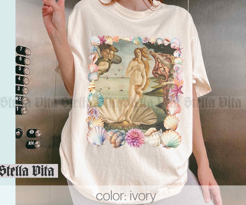 Puede incluir: Camiseta de color marfil con un estampado gr&aacute;fico de una pintura de estilo renacentista de una mujer de pie sobre una concha, rodeada de un borde floral de conchas. El texto "color: ivory" est&aacute; impreso en la parte inferior de la camiseta.