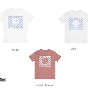 May include: Three t-shirts in different colors: white, ash, and heather mauve. Each shirt features a blue and white checkered pattern with a smiley face and the text "It's a beautiful day to shape behaviors".