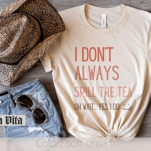 May include: A soft cream t-shirt with the text "I don't always spill the tea. Oh wait...yes I do." in pink lettering. The shirt is styled with denim shorts, a cowboy hat, and sunglasses.
