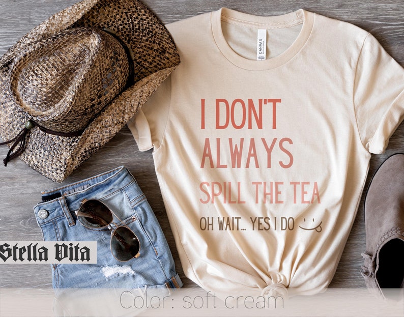 May include: A soft cream t-shirt with the text "I don't always spill the tea. Oh wait...yes I do." in pink lettering. The shirt is styled with denim shorts, a cowboy hat, and sunglasses.