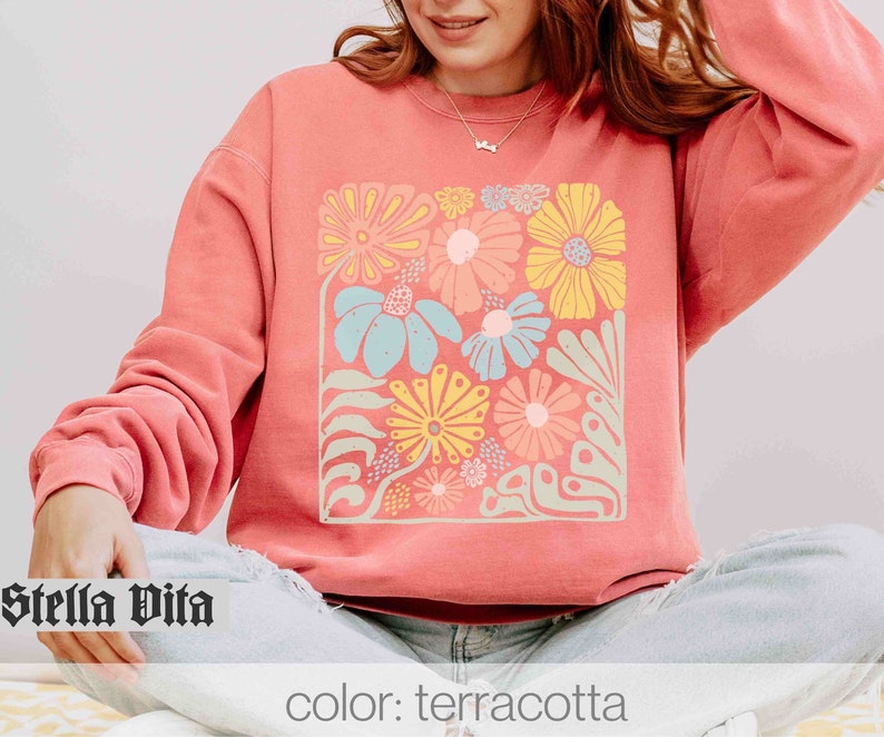 Peut inclure: Un sweat-shirt de couleur terre cuite avec un imprim&eacute; floral repr&eacute;sentant une vari&eacute;t&eacute; de fleurs color&eacute;es dans un motif carr&eacute;. Le texte "Stella Vita" est visible dans le coin inf&eacute;rieur gauche de l'image.