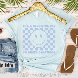 May include: A light blue t-shirt with a checkered pattern and a smiley face with hearts for eyes. The text "It's a beautiful day to shape behaviors" is printed on the shirt.