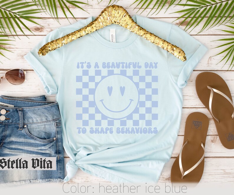 May include: A light blue t-shirt with a checkered pattern and a smiley face with hearts for eyes. The text "It's a beautiful day to shape behaviors" is printed on the shirt.