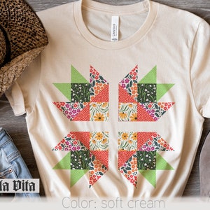 May include: A soft cream t-shirt with a floral patchwork design. The design features four squares with a green, red, and orange color scheme. The text "Stella Vita" is visible in the bottom left corner of the image.