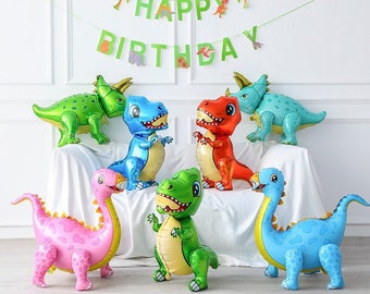 May include: A set of seven dinosaur-shaped foil balloons in various colors, including green, blue, red, pink, and yellow. The balloons are arranged on a white surface with a green and white banner that reads "Happy Birthday".