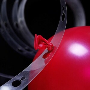 May include: A clear plastic balloon arch strip with holes for tying balloons. The strip is curved and has a red balloon tied to it.