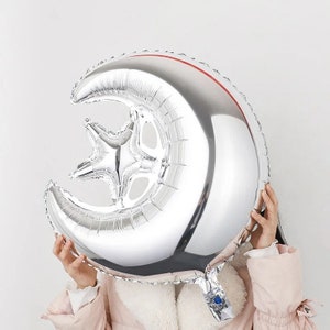 May include: A silver foil balloon shaped like a crescent moon with a star inside. The balloon is partially inflated and held in front of a person's face.