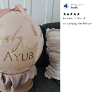 Może przedstawiać: Duży, przezroczysty balon o średnicy 61 cm z jasnor&oacute;żowym, błyszczącym wykończeniem. Balon jest ozdobiony złotą wstążką i ma napis "Baby AYUB" w złotym kolorze.