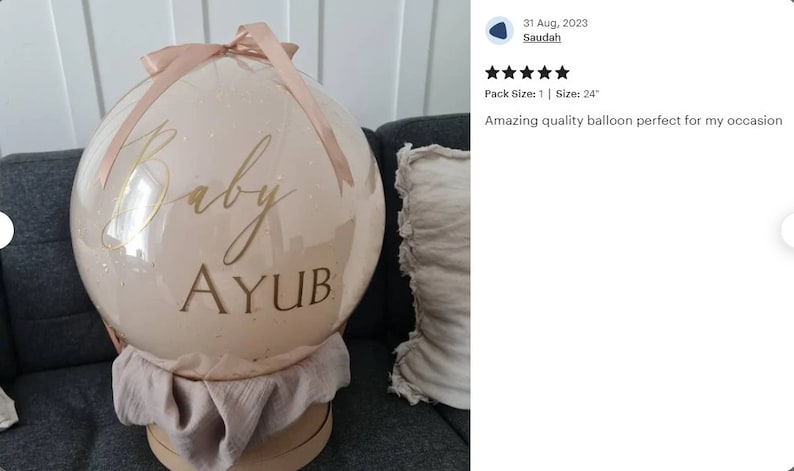 Może przedstawiać: Duży, przezroczysty balon o średnicy 61 cm z jasnor&oacute;żowym, błyszczącym wykończeniem. Balon jest ozdobiony złotą wstążką i ma napis "Baby AYUB" w złotym kolorze.