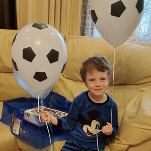 May include: A young boy wearing a blue and white striped pajamas with a Mickey Mouse graphic is sitting on a couch. He is holding a white and black soccer ball balloon. There is another soccer ball balloon behind him.