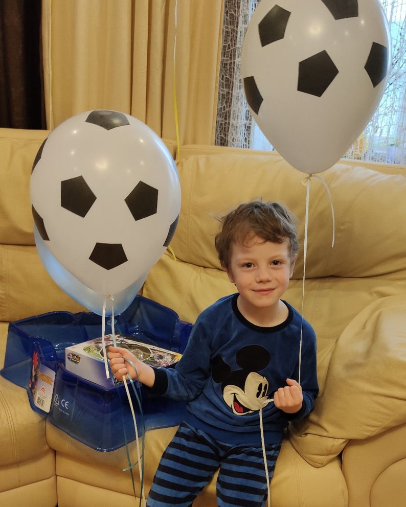 May include: A young boy wearing a blue and white striped pajamas with a Mickey Mouse graphic is sitting on a couch. He is holding a white and black soccer ball balloon. There is another soccer ball balloon behind him.