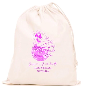 Könnte beinhalten: Ein cremefarbener Kordelzugbeutel mit einer rosa Grafik. Die Grafik zeigt eine Cowgirl auf einer Discokugel mit dem Text "Let's Go Girls" und "Jessica's Bachelorette Las Vegas, Nevada."