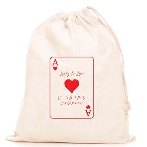 Könnte beinhalten: Ein cremefarbener Kordelzugbeutel mit einem roten Pik-Ass-Spielkarten-Design. Die Karte trägt den Text "Lucky In Love" und "Demi's Bach Party Las Vegas 2025". Die Tasche ist aus einem weichen, strukturierten Stoff gefertigt.