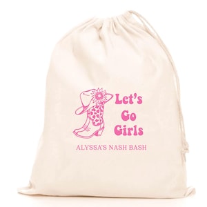 Könnte beinhalten: Ein cremefarbener Kordelzugbeutel mit einer rosa Grafik eines Cowboy-Stiefels und -Huts, verziert mit einer Sonnenblume. Der Text "Let's Go Girls" und "ALYSSA'S NASH BASH" sind ebenfalls in Rosa.