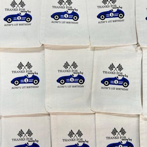 May include: A collection of off-white fabric drawstring bags, each printed with a blue race car graphic, two checkered flags, and the text: THANKS FOR racing by ALTAY'S 1ST BIRTHDAY. The design features a number one on the car body.