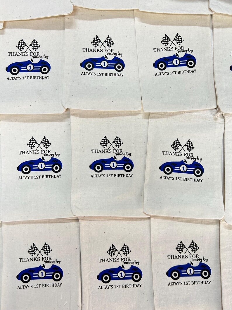 May include: A collection of off-white fabric drawstring bags, each printed with a blue race car graphic, two checkered flags, and the text: THANKS FOR racing by ALTAY'S 1ST BIRTHDAY. The design features a number one on the car body.