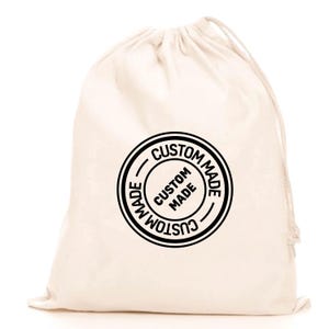 May include: A cream-colored drawstring bag with a black circular stamp that reads "CUSTOM MADE" in a bold, sans-serif font. The bag is made of a soft, natural fabric and has a simple, minimalist design.