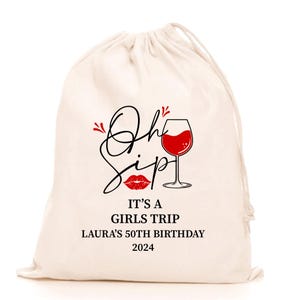 Op de afbeelding: Een crèmekleurige trekkoordtas met de tekst "Oh Sip" in zwart schrift, een rode lippenstiftafbeelding en een wijnglas gevuld met rode vloeistof. De tekst "IT'S A GIRLS TRIP LAURA'S 50TH BIRTHDAY 2024" staat ook op de tas.
