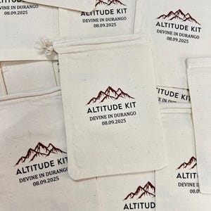 May include: A collection of off-white cotton drawstring pouches featuring a printed design in dark brown. The graphic includes a mountain range icon above the text ALTITUDE KIT, DEVINE IN DURANGO, and the date 08.09.2025 centred on each bag.