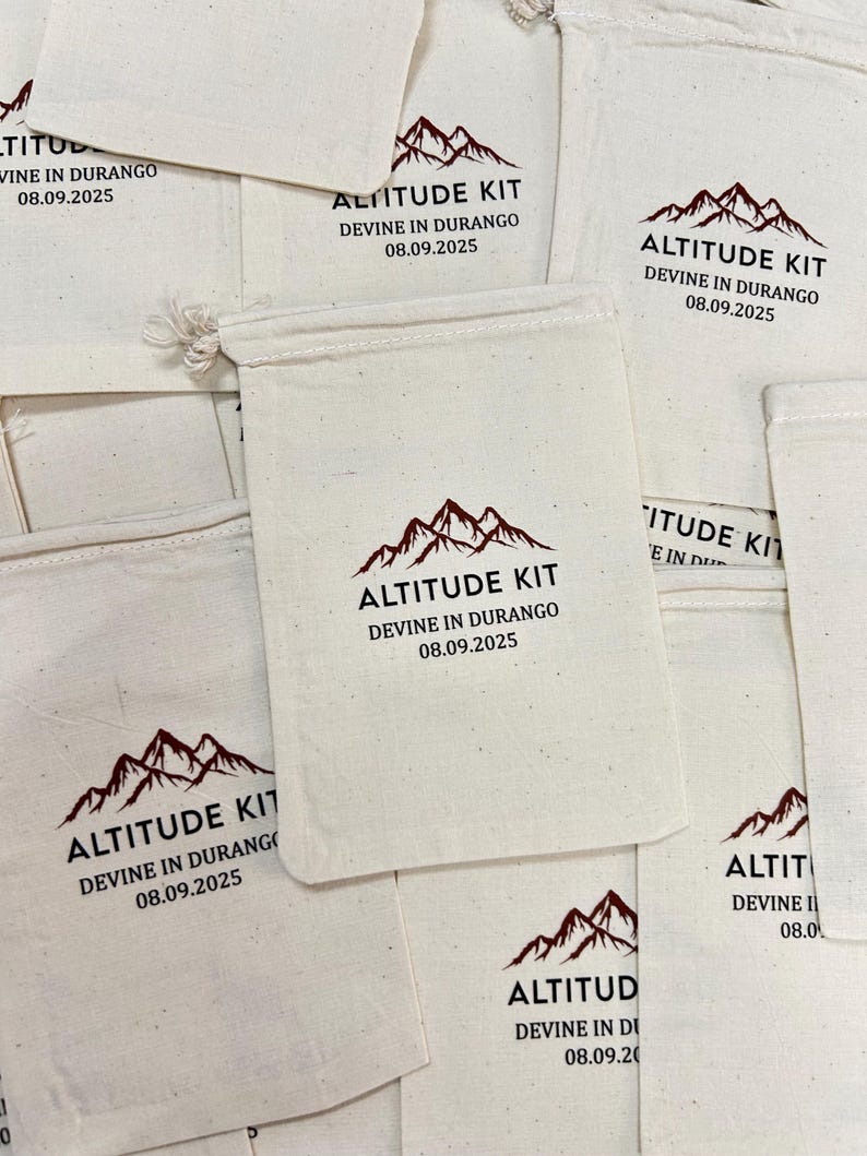 May include: A collection of off-white cotton drawstring pouches featuring a printed design in dark brown. The graphic includes a mountain range icon above the text ALTITUDE KIT, DEVINE IN DURANGO, and the date 08.09.2025 centred on each bag.
