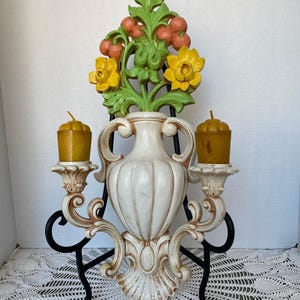 May include: A decorative wall sconce featuring a white vase with yellow and orange floral accents. Two candle holders with gold candles are attached to the vase. The sconce is supported by a black metal stand.